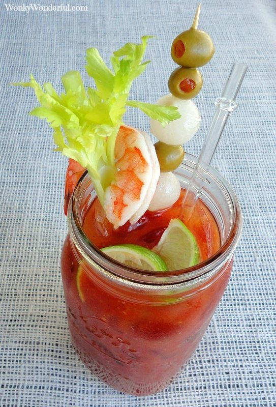 Bloody Mary cocktail in large clear glass jar topped with celery, shrimp and olives