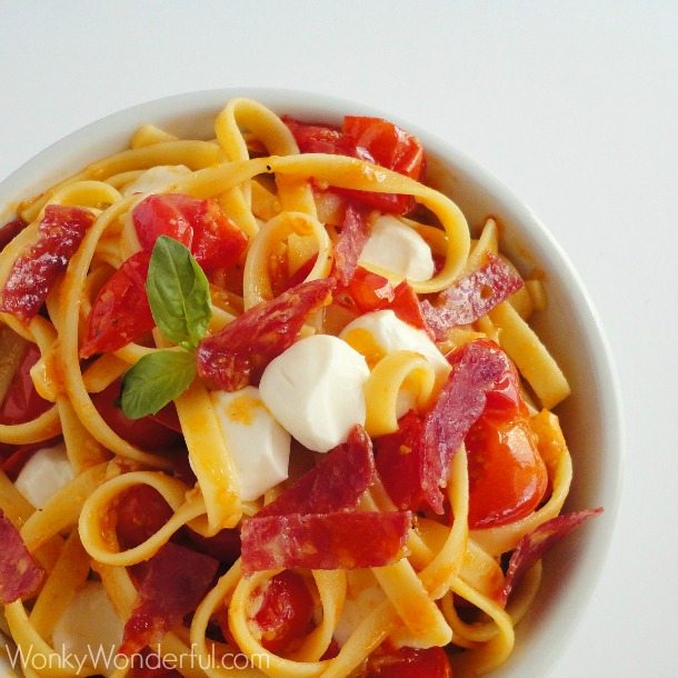 above view of pasta, tomatoes, mozzarella, salami and basil in white bowl
