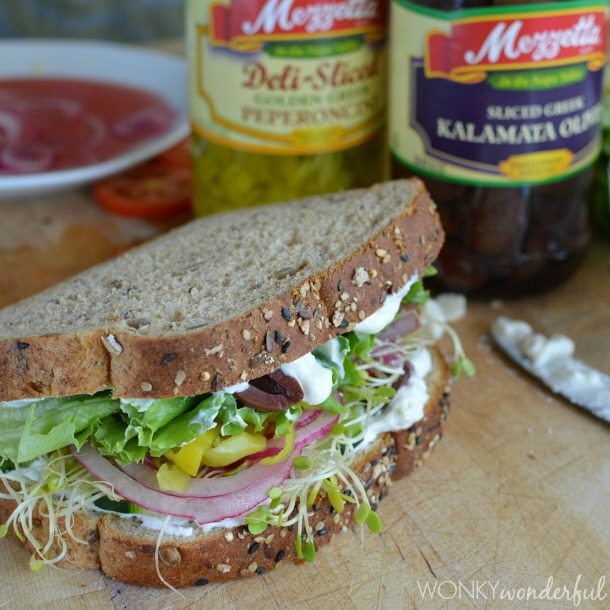 prepared sandwich on cutting board next to jars or pepperoncinis and olives
