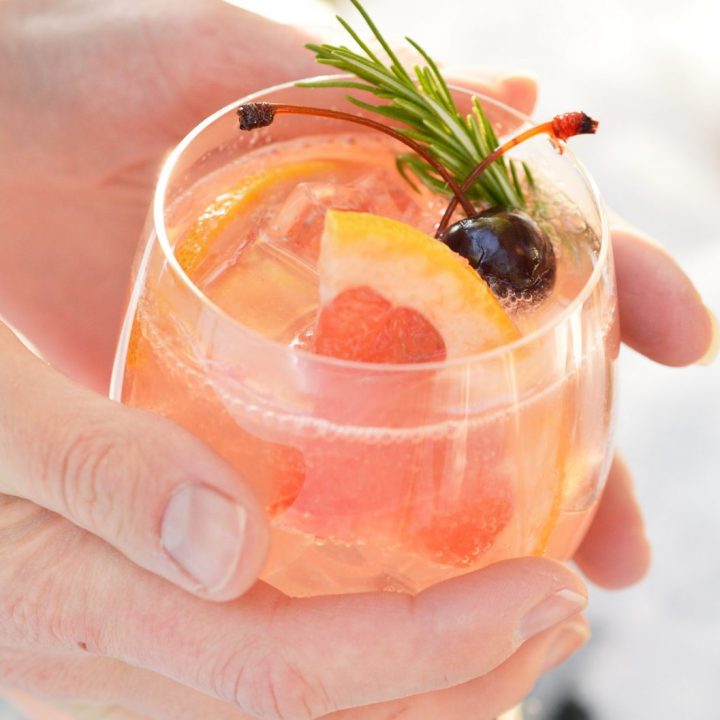 two hands holding glass filled with fizzy cocktail, grapefruit and cherries