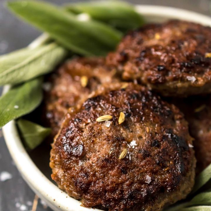 close up of homemade breakfast sausage recipe finished. browned sausage patties topped with fennel seeds and green sage in the background