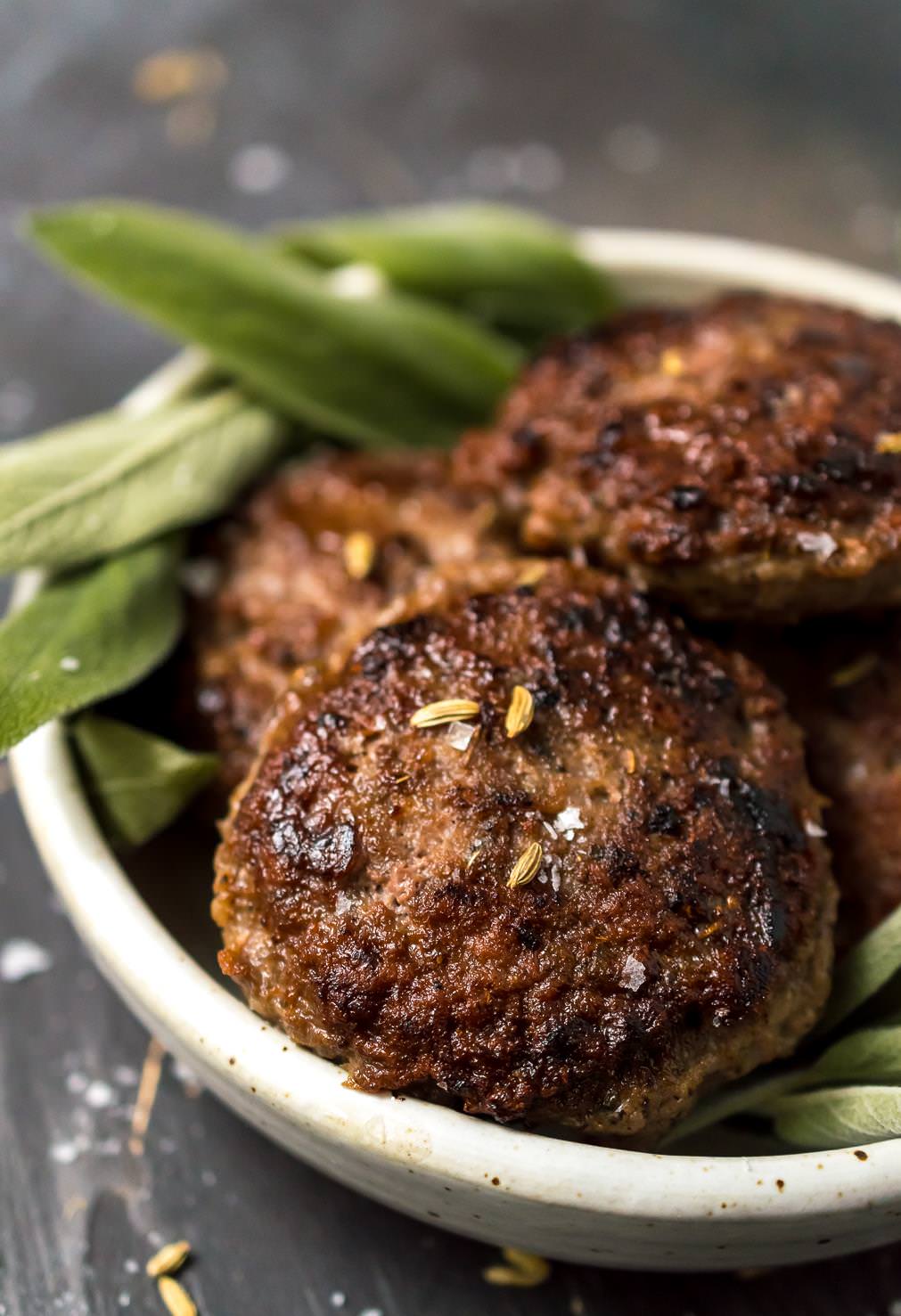close up of homemade breakfast sausage recipe finished. browned sausage patties topped with fennel seeds and green sage in the background