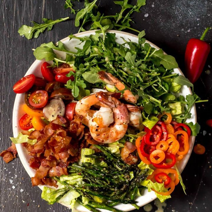 overhead photo of a messy salad topped with roasted asparagus shrimp bacon tomatoes mini peppers and lemon pepper dressing, white bowl black background
