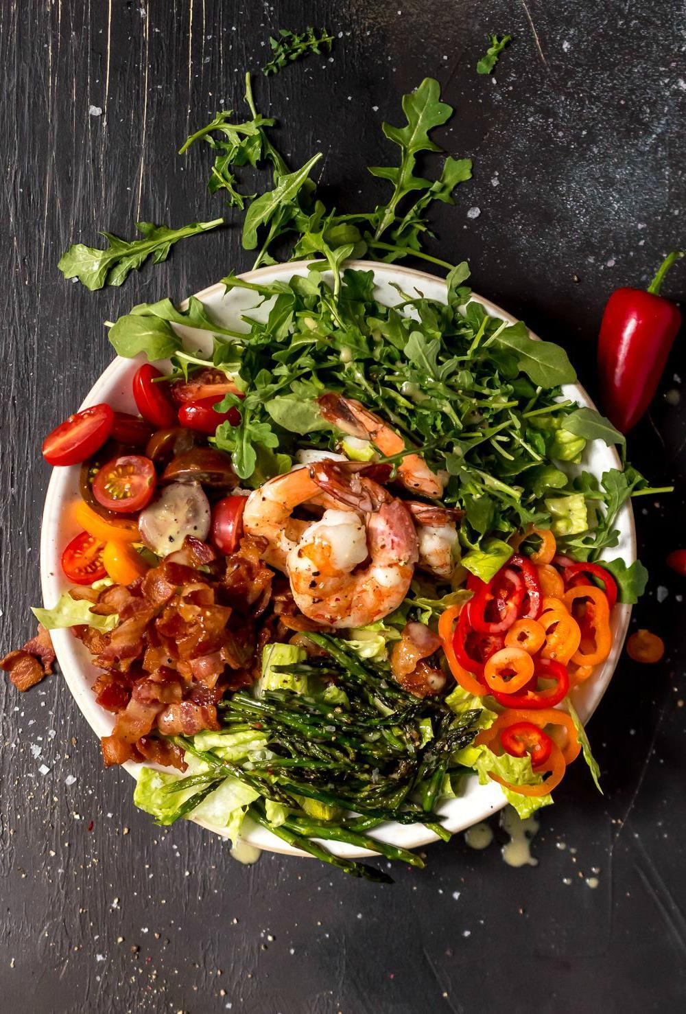 overhead photo of a messy salad topped with roasted asparagus shrimp bacon tomatoes mini peppers and lemon pepper dressing, white bowl black background