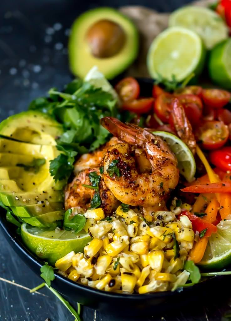 close up of spicy shrimp coated in seasoning with bright yellow roasted corn kernels in front and a sliced avocado on the side