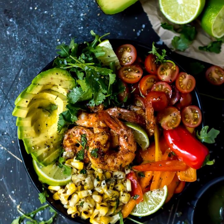 overhead shot of spicy shrimp bowl filled with fresh colorful ingredients, bluish-black background with avocados and limes on the side