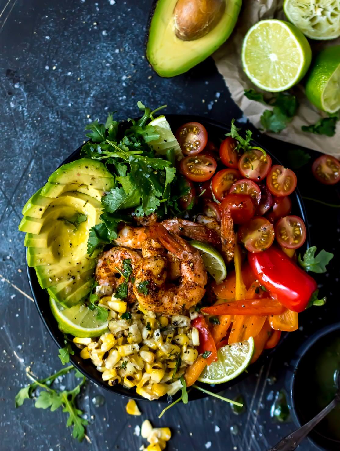 overhead shot of spicy shrimp bowl filled with fresh colorful ingredients, bluish-black background with avocados and limes on the side
