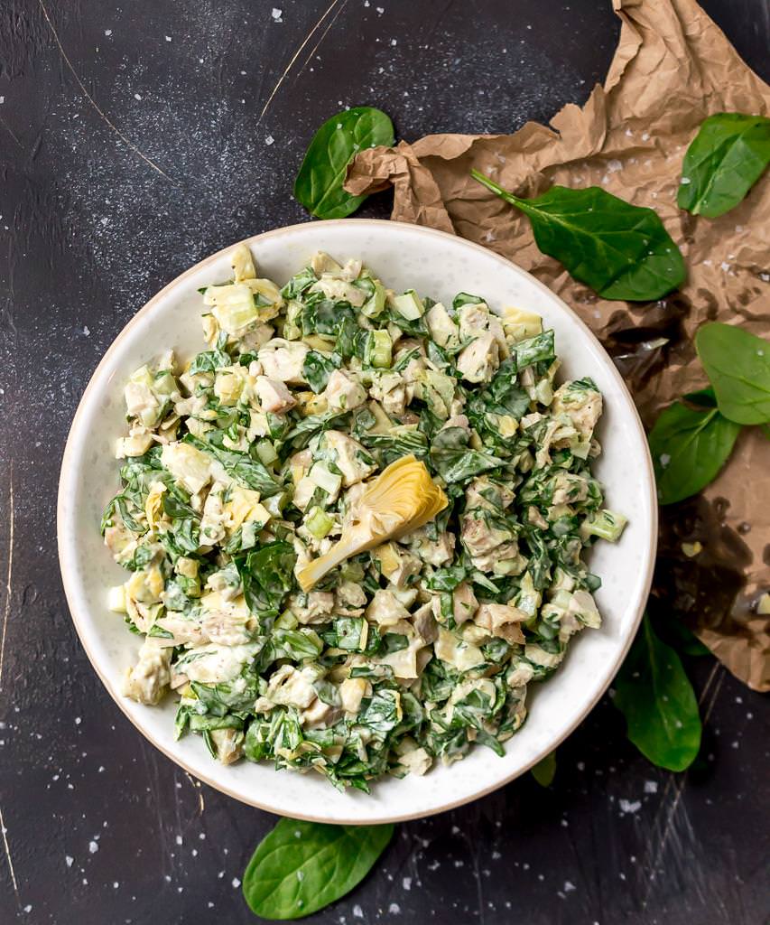 overhead photo of spinach artichoke chicken salad in white bowl. brown wrinkled paper and fresh spinach leaves on the side
