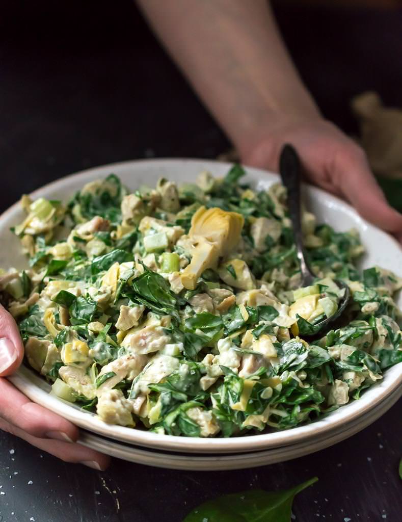 two hands carrying white bowl filled with spinach artichoke chicken salad for serving