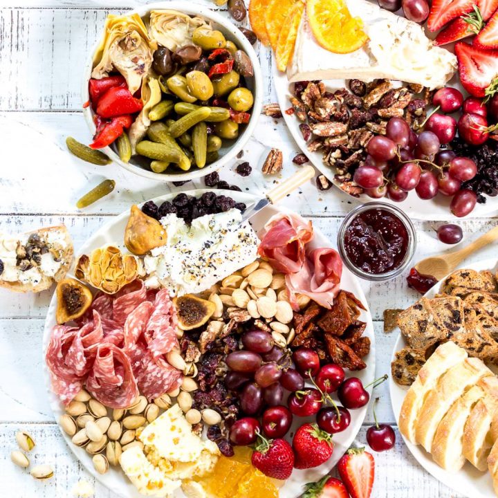 over head photo of cheese board spread on white background. cheese, salami, fresh fruit, dried fruit, nuts, olives, honey and more