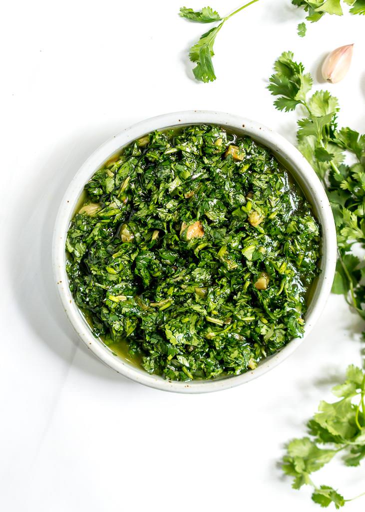 over head image of bright green roasted garlic cilantro chimichurri sauce with cilantro and garlic on the side