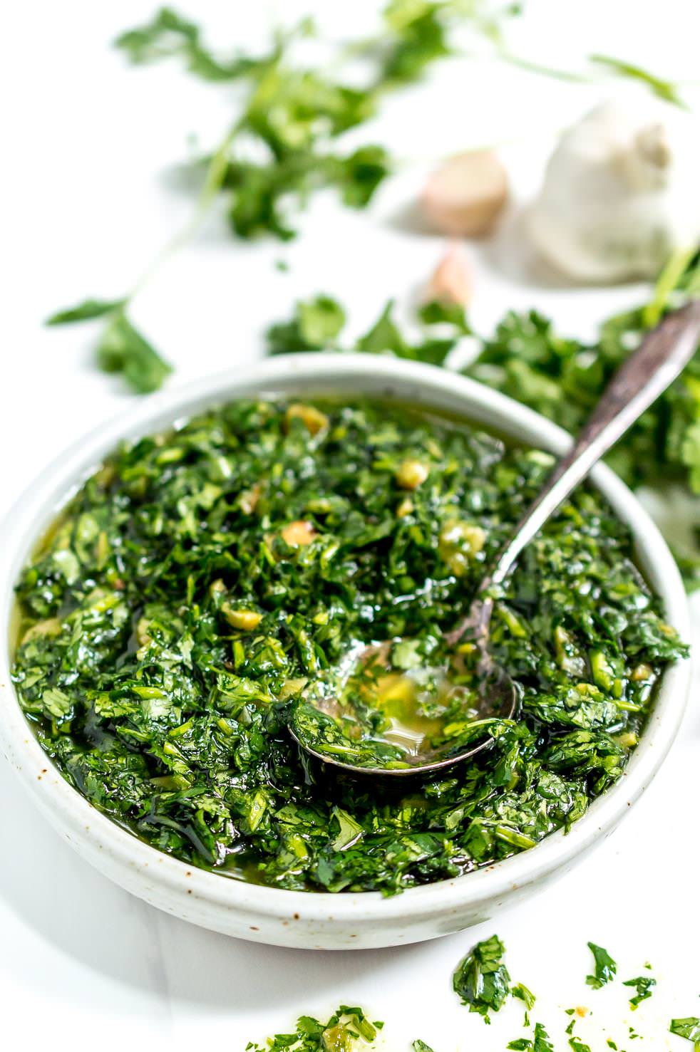 close up photo of white bowl filled with roasted garlic chimichurri sauce and a serving spoon, fresh garlic and cilantro in the background