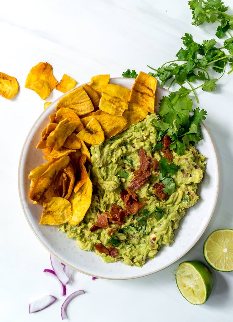 over head photo of bacon guac recipe mixed together in a white bowl with plantain chips on the side