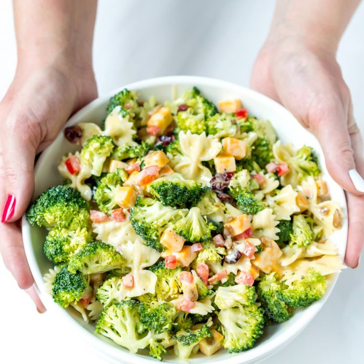 two hands holding white bowl filled with broccoli pasta salad