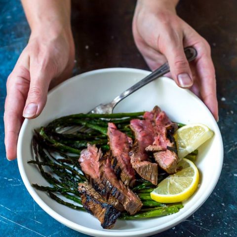 flank steak over roasted asparagus with lemons in white bowl held by two hands
