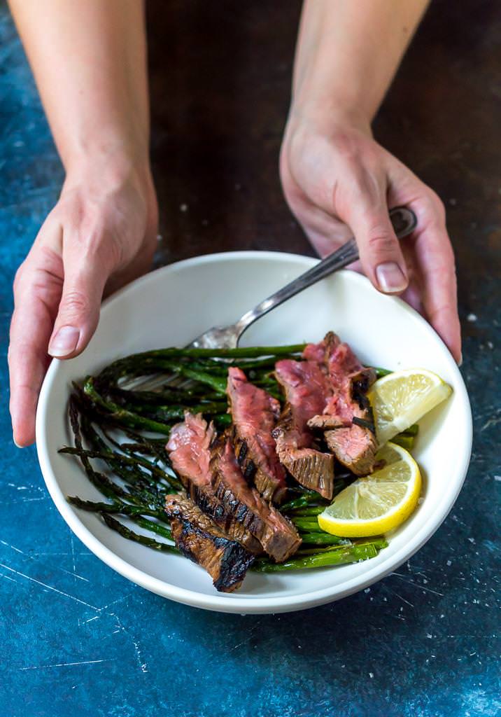 flank steak recipe over roasted asparagus with lemons in white bowl held by two hands