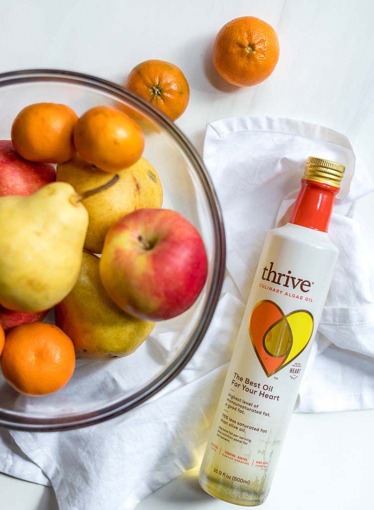thrive oil next to a glass bowl filled with apples, pears and mandarins