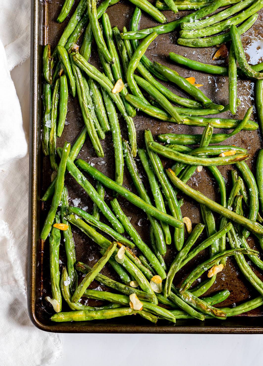 roasted fresh green beans with slices of golden garlic in a dark metal baking sheet