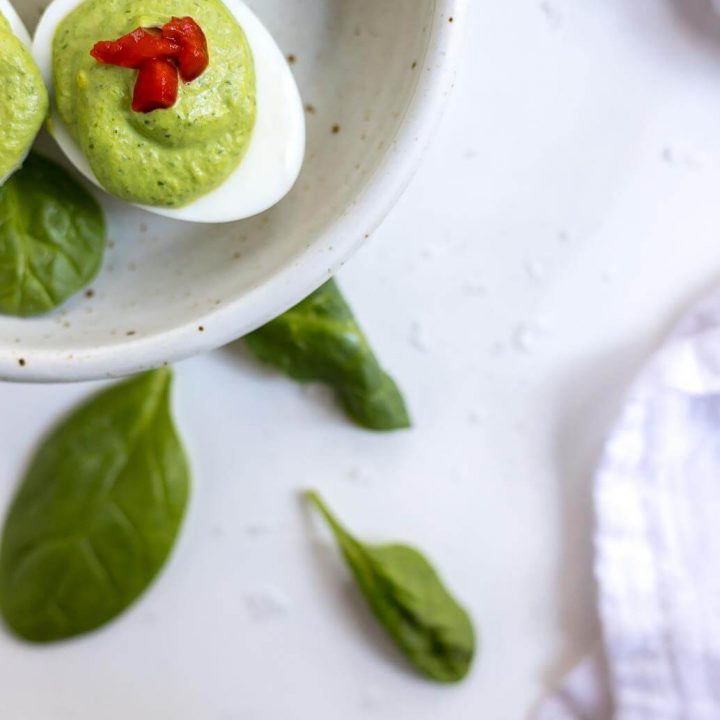 green deviled egg recipe topped with red pimentos in white dish with spinach on the side