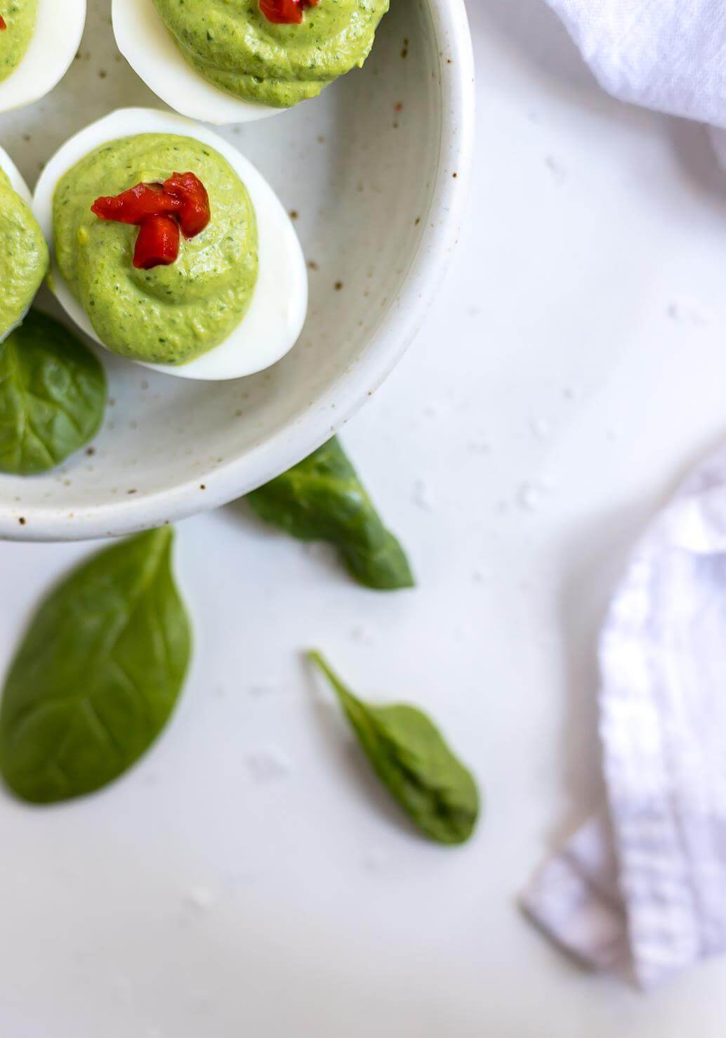 green deviled egg recipe topped with red pimentos in white dish with spinach on the side