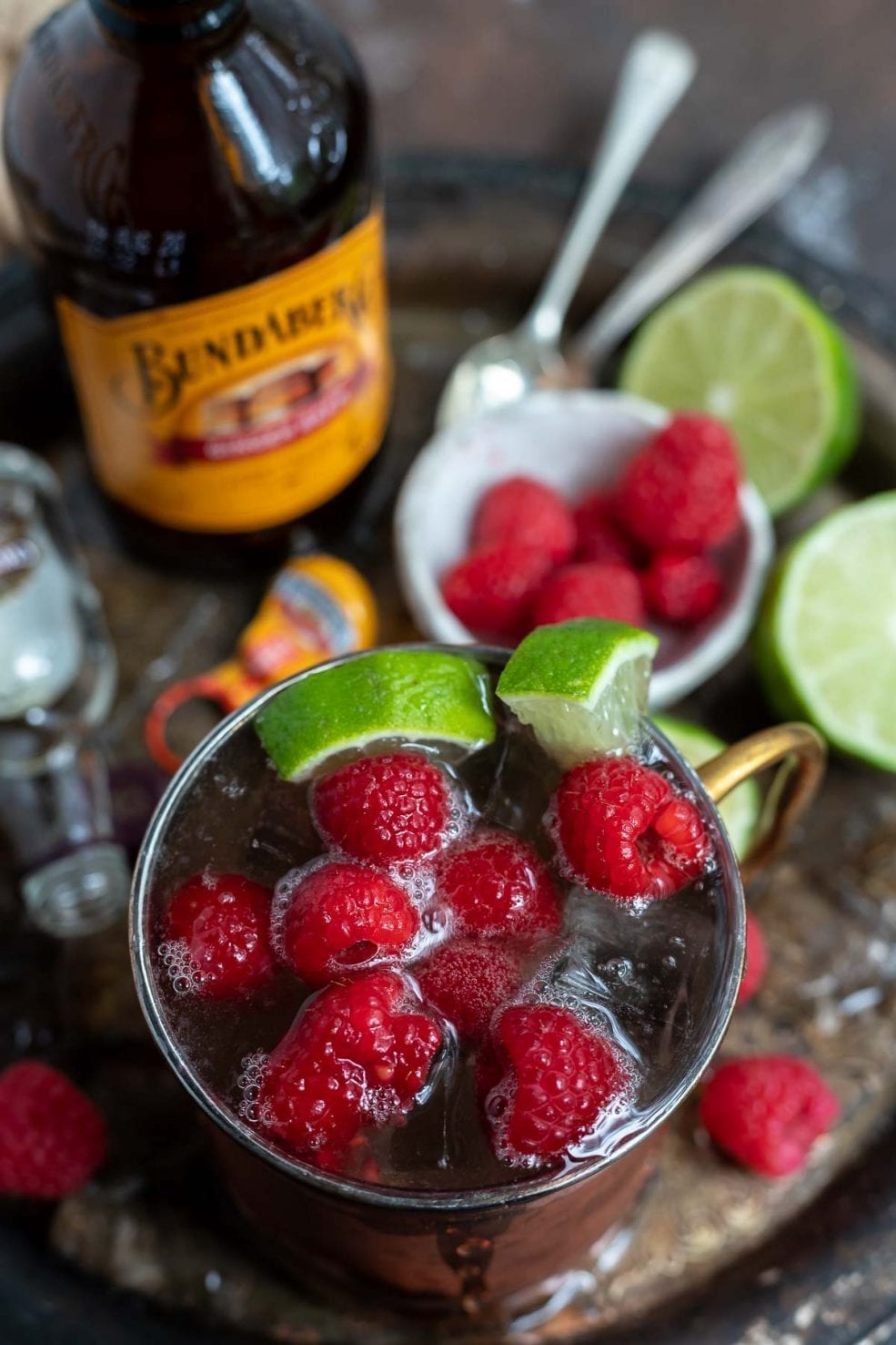 drink in copper mug next to fresh raspberries, limes and ginger beer bottle