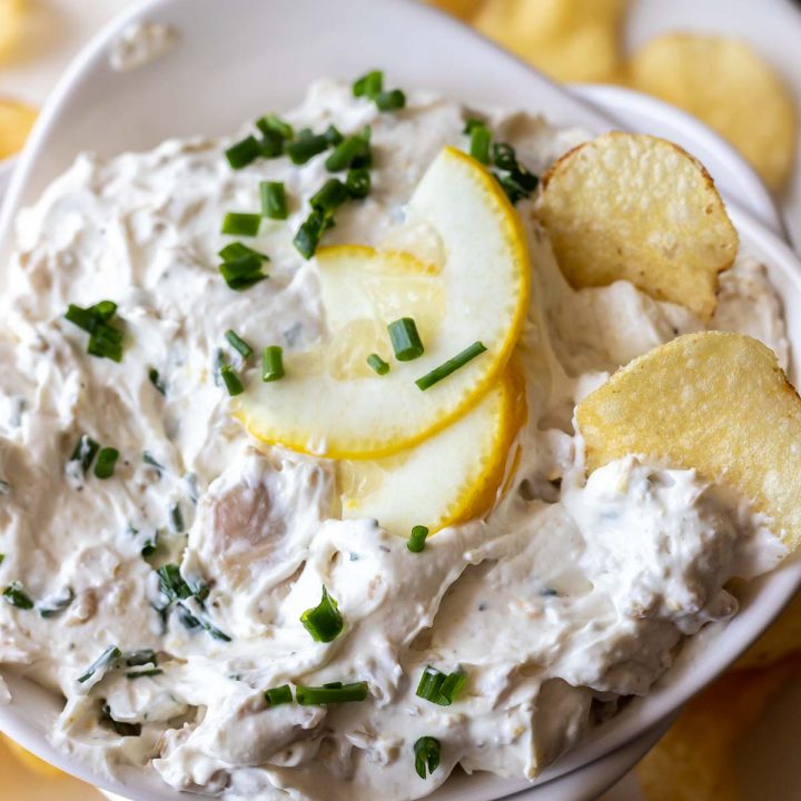 creamy dip topped with lemon slices and chopped chives in white bowl next to potato chips